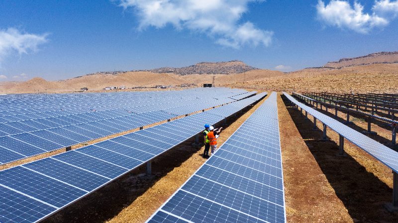 Panel sıralarının arasında baret ve reflektörlü yelek giymiş iki saha çalışanı, arkada Mardin tepeleri
