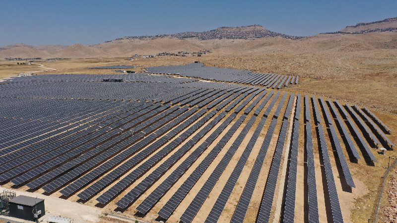 Santral sahasının alçak açıdan havadan görünümü: paralel panel sıraları, servis yolları ve önde bir invertör kabini